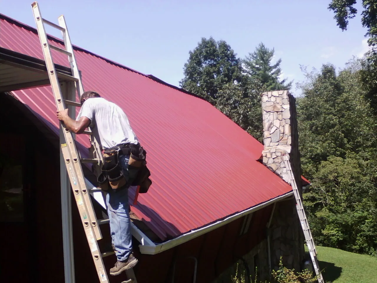 Roofer on a ladder beside a red metal roof and stone chimney — Soffit & Fascia Repair by Roofing Brothers in Thiells